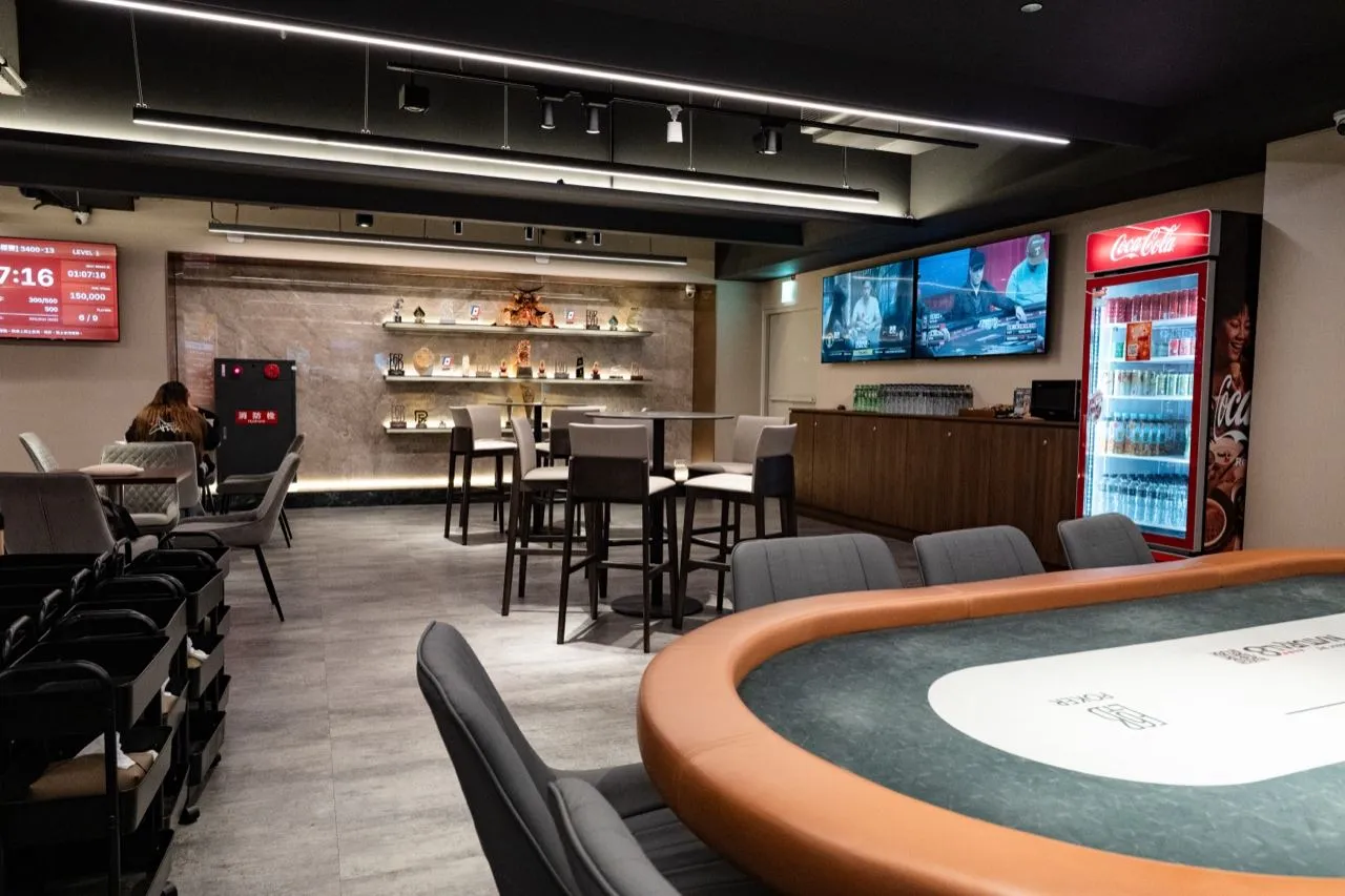 6BET 的吧檯區，高腳桌椅前方是展示架與飲料冰箱，左側牆上的螢幕顯示賽事時鐘，前景是一張牌桌的邊角