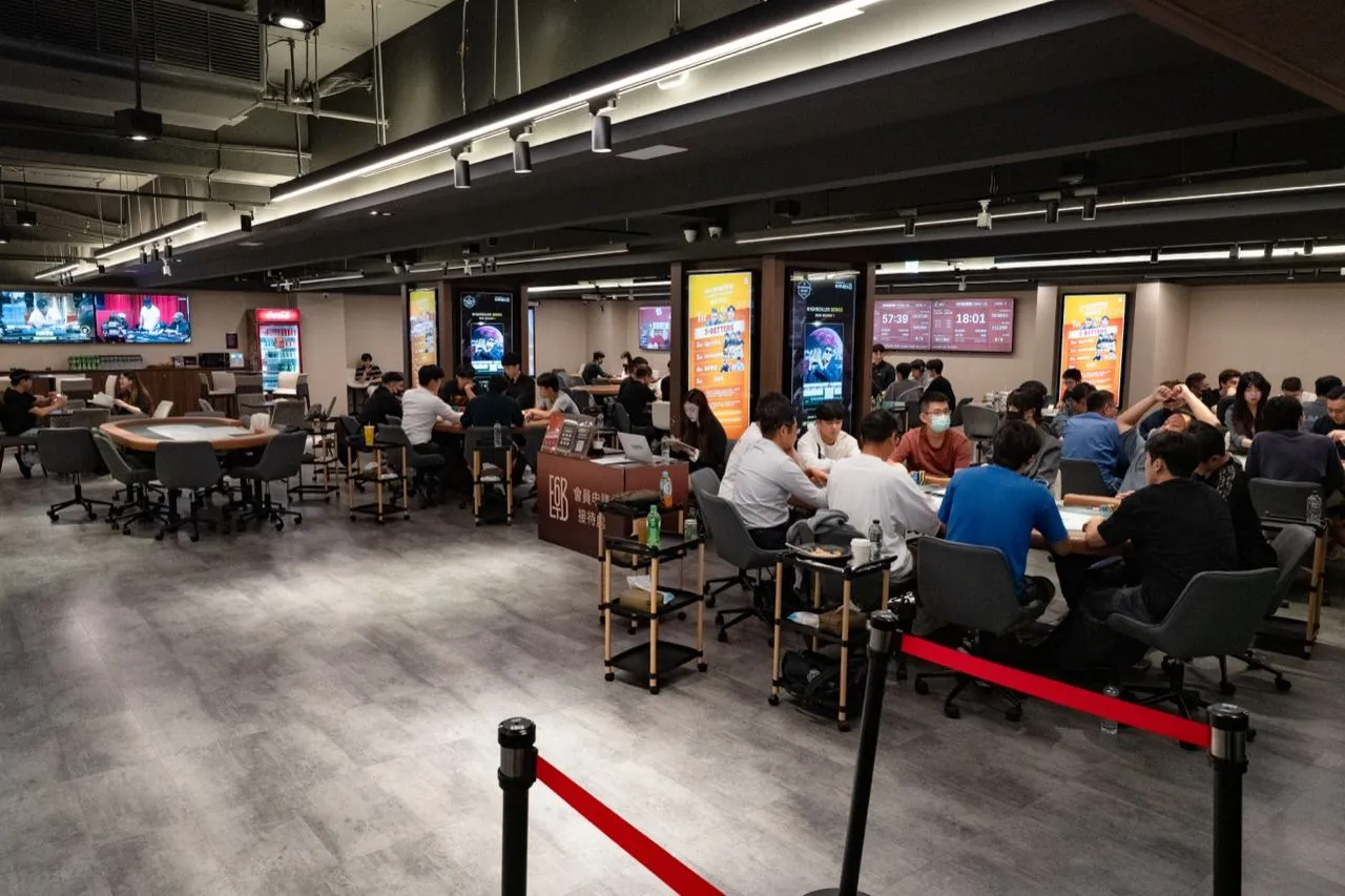 6BET 賽場全景，紅色圍欄外是多張坐滿選手的牌桌，天花板下方一排燈箱顯示各場賽事資訊，左側是尚未開桌的空桌
