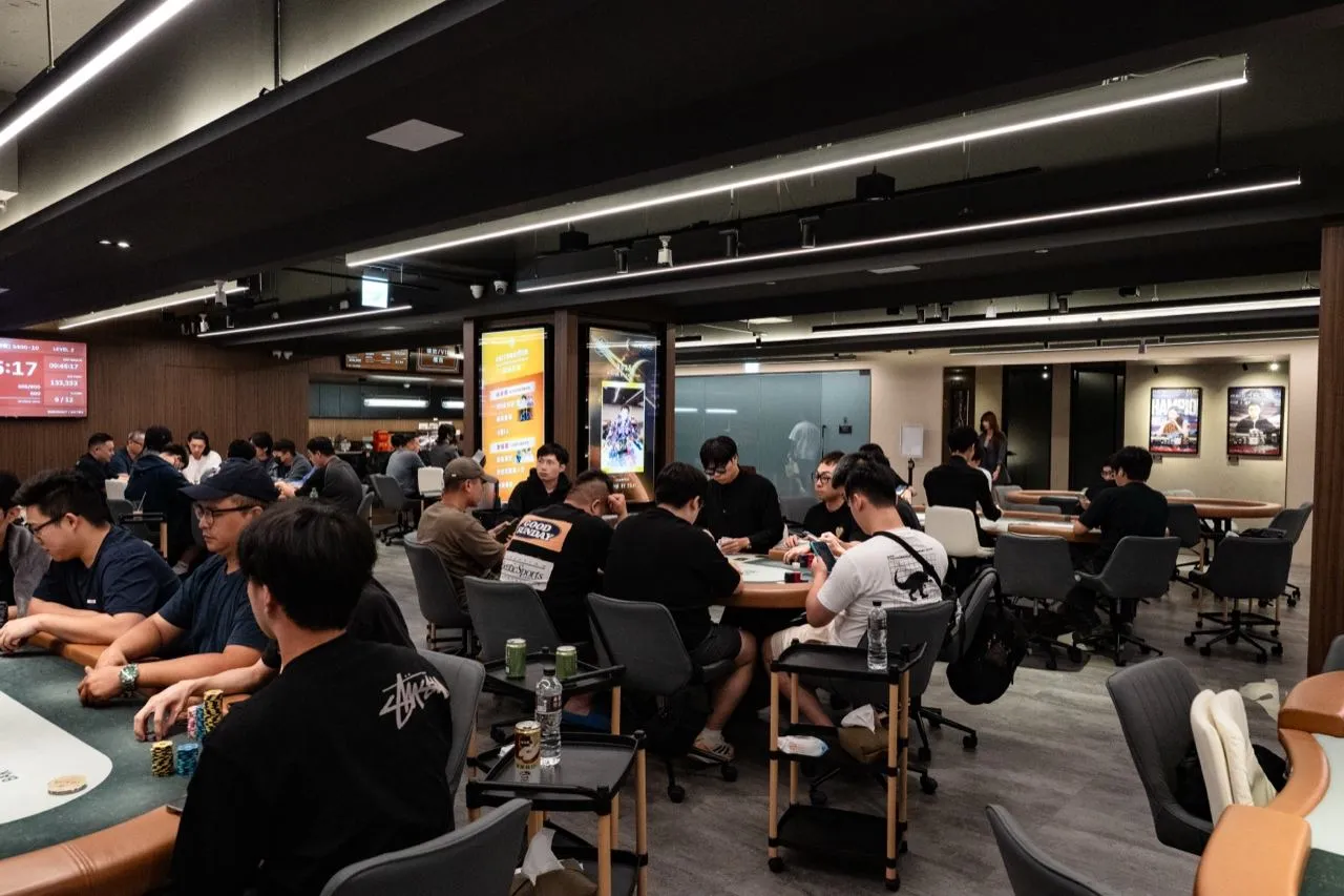 從側面看 6BET 賽場，前景幾張牌桌有選手入座，牆邊是賽程燈箱，遠處可見包廂區的玻璃門