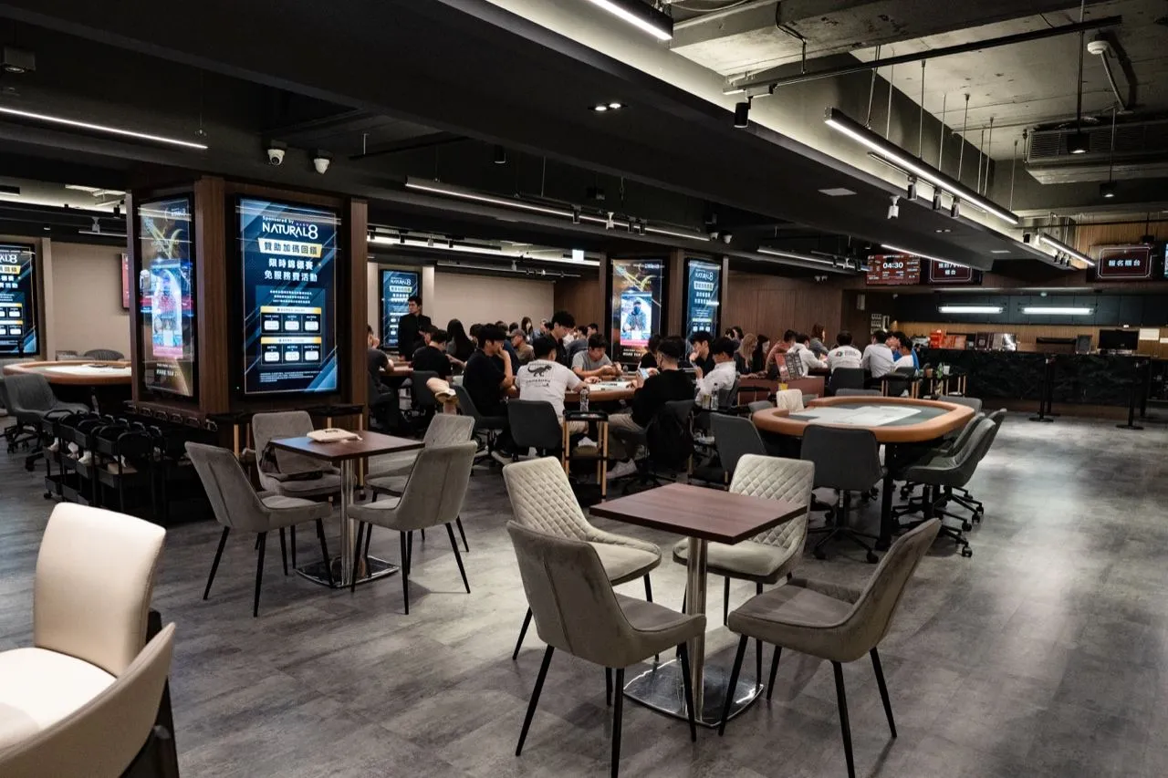 6BET 的休息用餐區，灰色與米色的餐椅配深色木紋小圓桌，後方是坐著選手的牌桌與賽程燈箱
