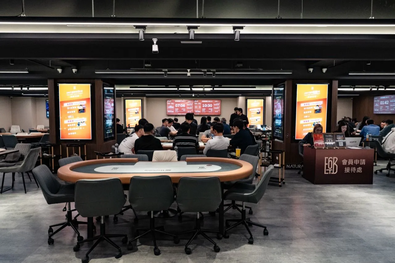 6BET 主賽場前排，一張空牌桌配灰色座椅，後方各桌坐著正在比賽的選手，兩側的直立燈箱顯示賽程，右側是深紅色的會員申請接待處