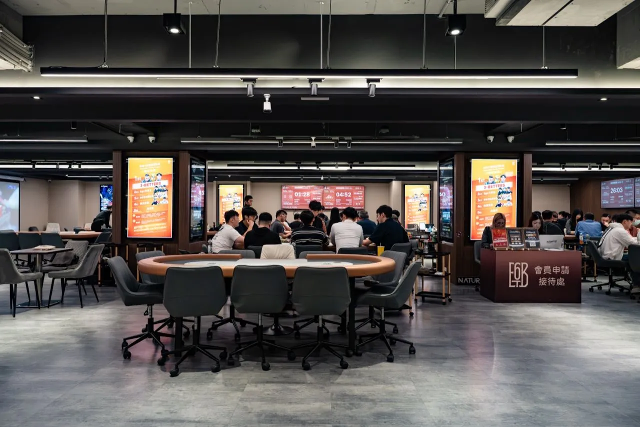 6BET 主賽場全景，前方一張淺綠桌面的空牌桌，後方多桌選手正在進行賽事，右側是會員申請接待處與賽程燈箱