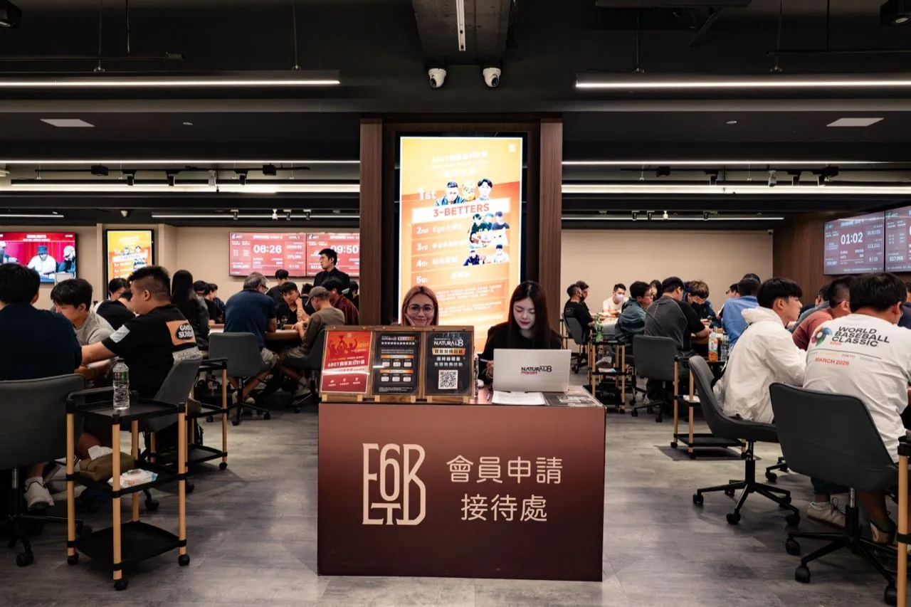 6BET 會員申請接待處特寫，深紅色櫃台上放著立牌與 QR code，工作人員坐在櫃台後方，背景是多桌進行中的賽事