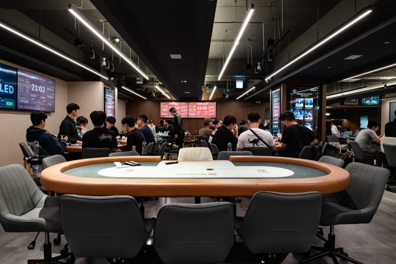 6BET 賽場一景，前景是一張空的淺綠桌面牌桌，後方各桌坐滿選手，左上方螢幕顯示賽事時鐘與盲注級別