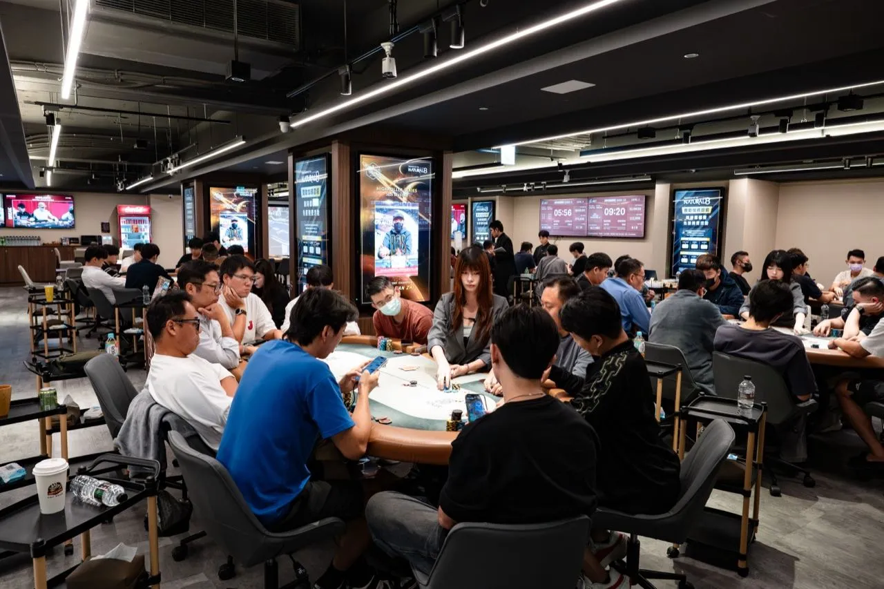 6BET 賽場裡多張牌桌坐滿選手，荷官站在桌前發牌，牆上的燈箱與螢幕顯示各場賽事的時鐘與盲注