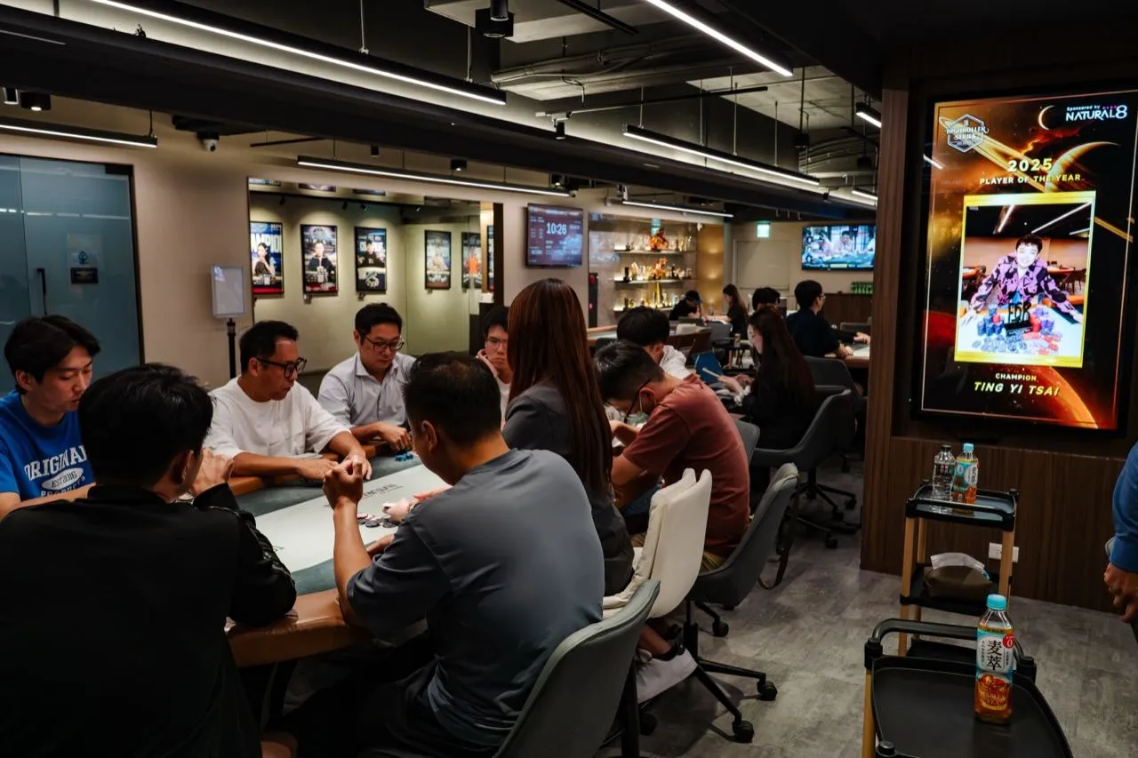 6BET 賽場一角，選手圍著牌桌進行賽事，右側牆上的燈箱展示 2025 年度最佳選手海報，後方牆面掛著歷屆冠軍照片